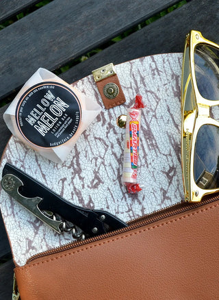 A travel-friendly solid massage bar is shown spilling out of a purse with sunglasses, candy, and a corkscrew.